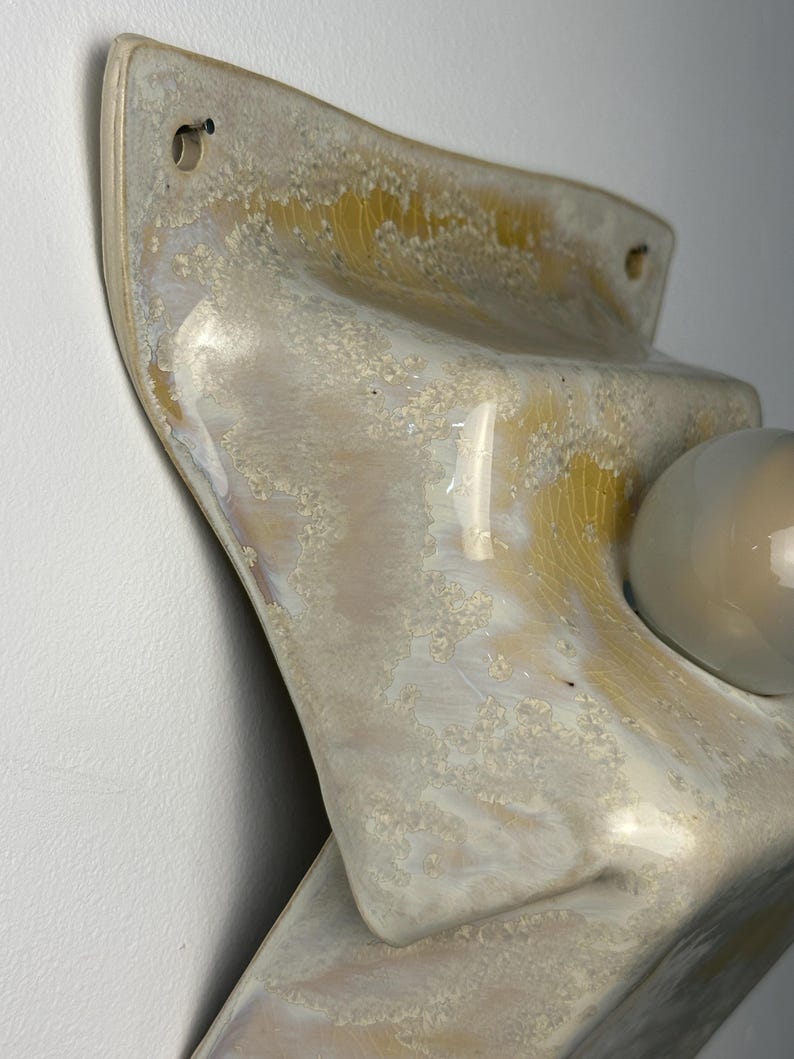 May include: A wall-mounted ceramic light fixture featuring a glossy, textured glaze in shades of cream, beige, and pale yellow. The surface has a crackled, crystalline appearance with a smooth, rounded white glass bulb protruding from the center.