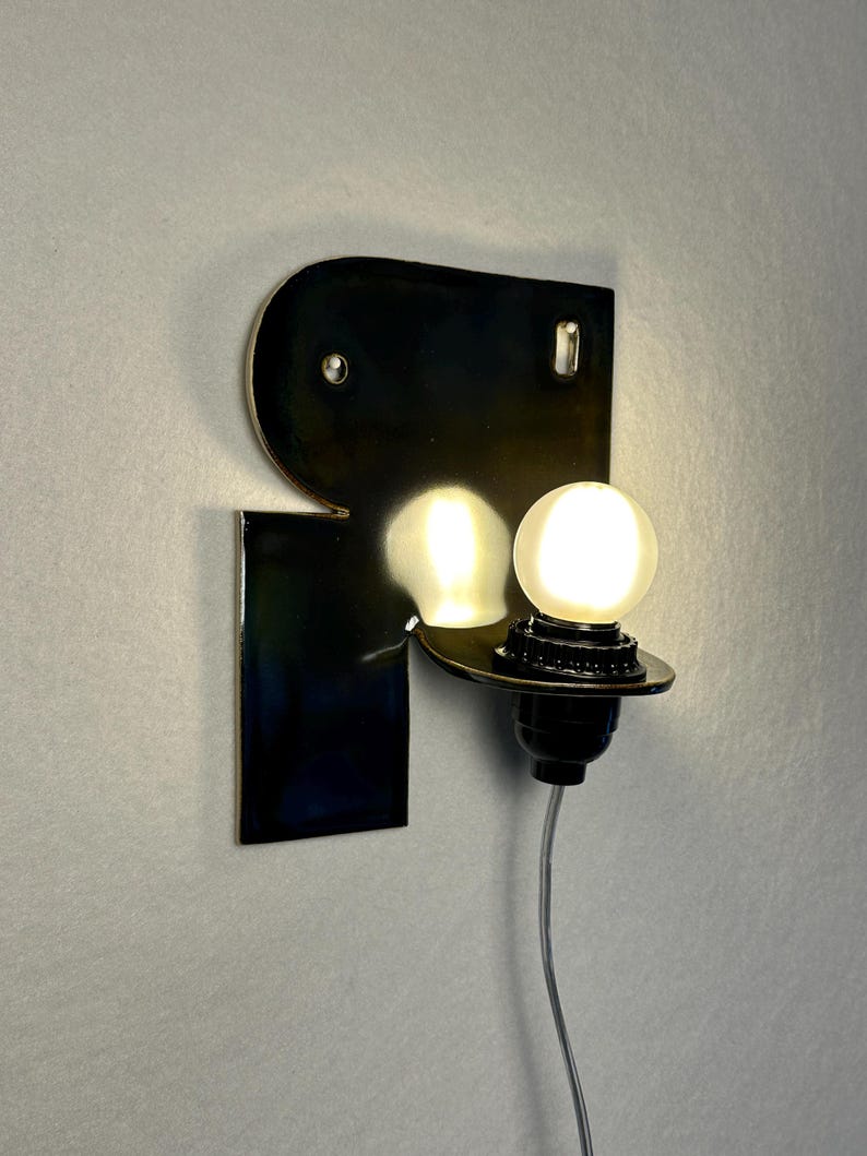 May include: A wall-mounted lamp featuring a glossy, dark, reflective metal backplate shaped like a stylised letter R. A single, round, glowing white light bulb is mounted on a black socket extending from the lower right side of the metal plate.