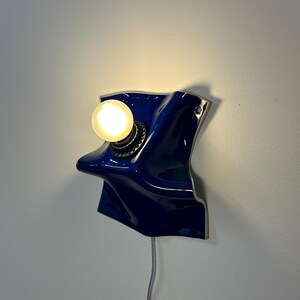 May include: A wall-mounted lamp featuring a glossy, deep blue, irregularly shaped ceramic base with a wavy, folded surface. A single round, warm-glowing light bulb is mounted on the left side of the blue fixture, which includes a thin power cord.