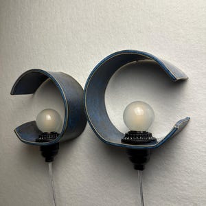 May include: Two wall-mounted lamps featuring curved, deep blue textured shades with light-colored edges. Each lamp has a black socket holding a round, frosted white light bulb and a clear power cord extending downward from the base.