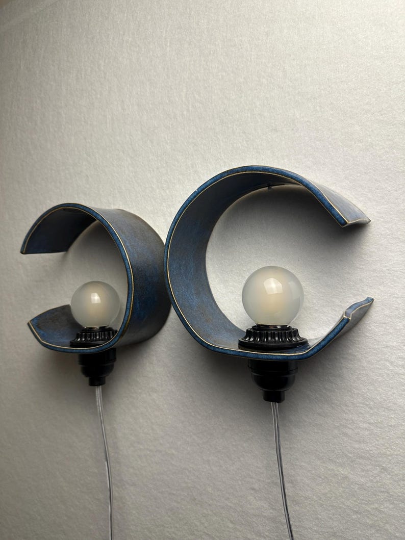 May include: Two wall-mounted lamps featuring curved, deep blue textured shades with light-colored edges. Each lamp has a black socket holding a round, frosted white light bulb and a clear power cord extending downward from the base.