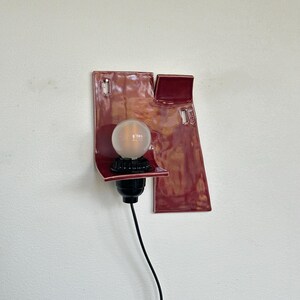 以下が含まれることがあります： ユニークな抽象的なデザインのモダンな壁掛け照明器具。器具は、電球ソケットと透明な丸い電球を備えた濃い赤色のセラミックベースを備えています。黒い電源コードが下に伸びています。