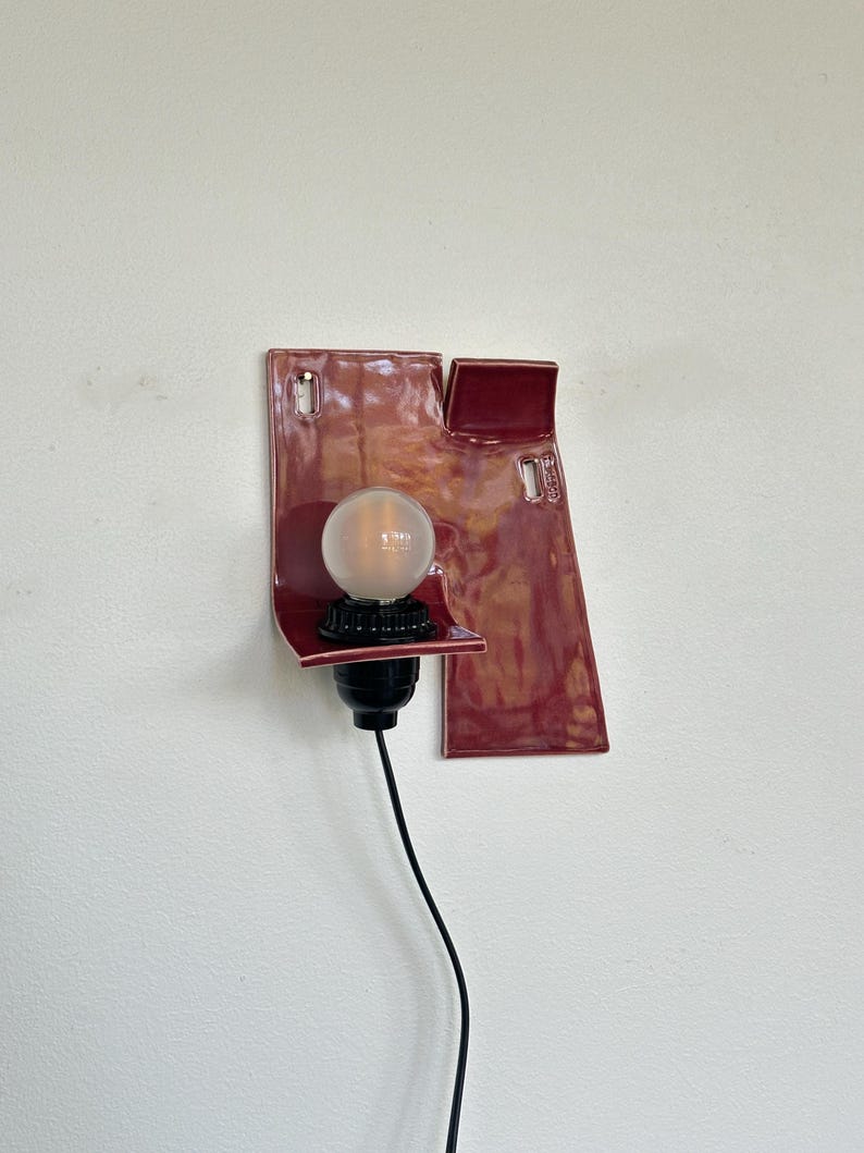以下が含まれることがあります： ユニークな抽象的なデザインのモダンな壁掛け照明器具。器具は、電球ソケットと透明な丸い電球を備えた濃い赤色のセラミックベースを備えています。黒い電源コードが下に伸びています。