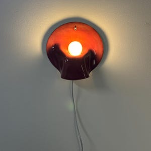 May include: A wall-mounted lamp featuring a circular, bowl-shaped shade with a glossy finish that transitions from a deep dark red at the bottom to a vibrant orange at the top. A single glowing white light bulb is centred within the shade.