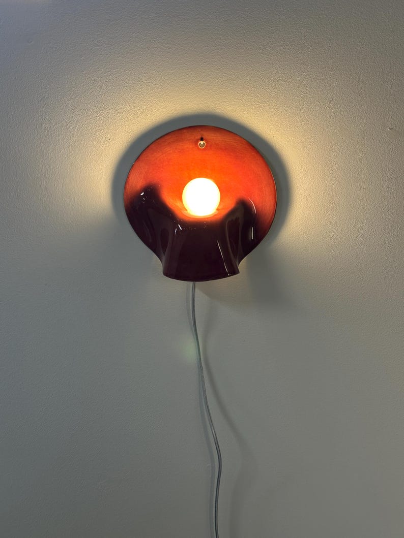 May include: A wall-mounted lamp featuring a circular, bowl-shaped shade with a glossy finish that transitions from a deep dark red at the bottom to a vibrant orange at the top. A single glowing white light bulb is centred within the shade.