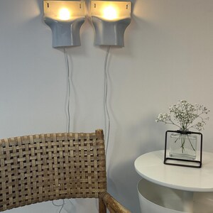 May include: Two light blue wall-mounted sconces with glowing bulbs, each featuring a rectangular top and a curved base. The lamps have white power cords hanging down. A woven chair and a small glass vase with white flowers are also visible.