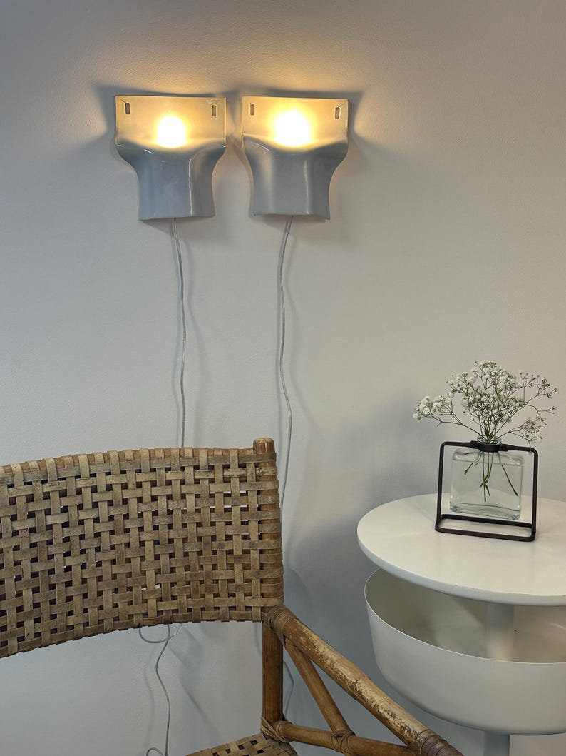 May include: Two light blue wall-mounted sconces with glowing bulbs, each featuring a rectangular top and a curved base. The lamps have white power cords hanging down. A woven chair and a small glass vase with white flowers are also visible.