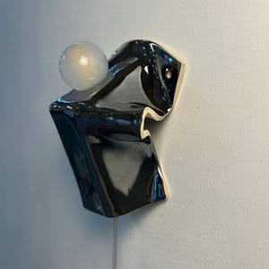 May include: A wall-mounted ceramic light fixture featuring a glossy black, folded, abstract form with a smooth finish. A single, spherical, frosted white glass light bulb is positioned on the upper section of the dark, sculptural ceramic base.