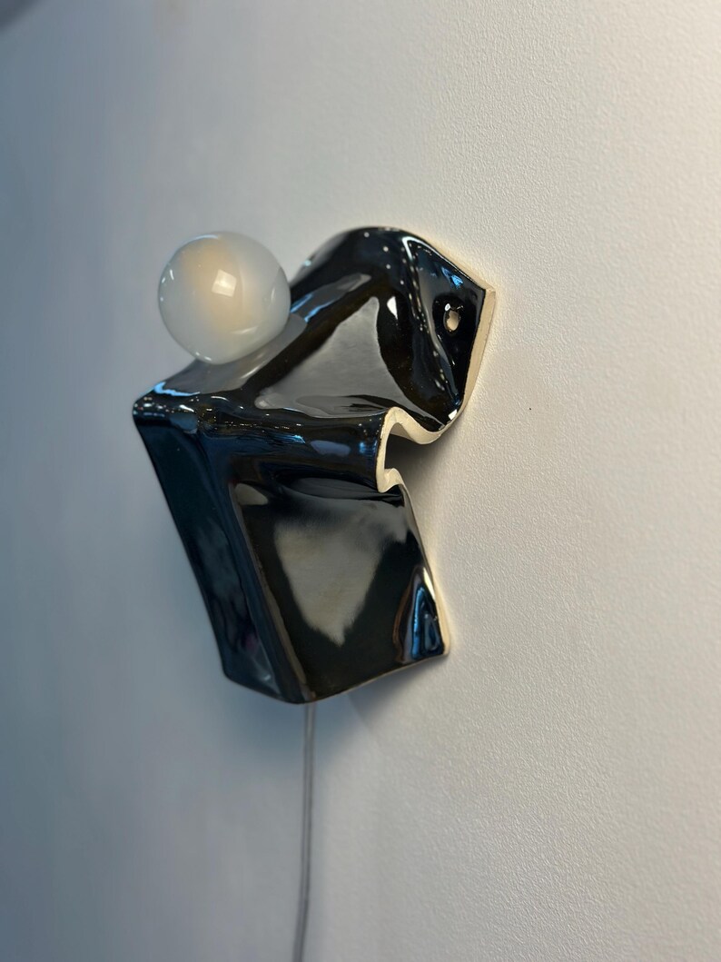May include: A wall-mounted ceramic light fixture featuring a glossy black, folded, abstract form with a smooth finish. A single, spherical, frosted white glass light bulb is positioned on the upper section of the dark, sculptural ceramic base.