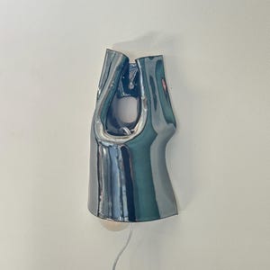 May include: A wall-mounted lamp with a glossy, dark metallic finish and an irregular, fluid shape. The fixture features a central cutout and a curved, reflective surface that catches light, with a thin, clear power cord extending from the bottom.
