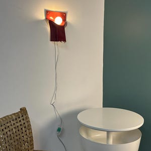 Pode incluir: Uma lumin&aacute;ria de parede com c&uacute;pula de couro vermelho escuro em design curvo e dobrado. A l&acirc;mpada emite uma luz quente a partir de uma l&acirc;mpada exposta. Um fio el&eacute;trico fino e transparente pende da lumin&aacute;ria com um pequeno interruptor.