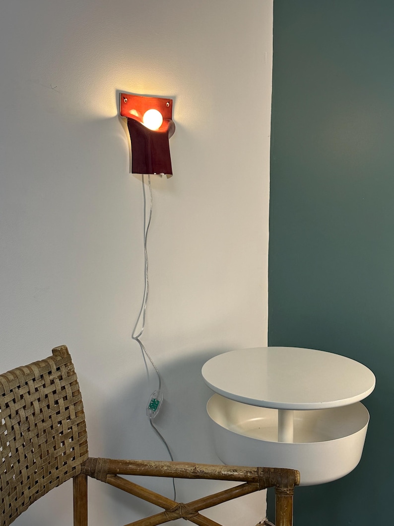 Pode incluir: Uma lumin&aacute;ria de parede com c&uacute;pula de couro vermelho escuro em design curvo e dobrado. A l&acirc;mpada emite uma luz quente a partir de uma l&acirc;mpada exposta. Um fio el&eacute;trico fino e transparente pende da lumin&aacute;ria com um pequeno interruptor.