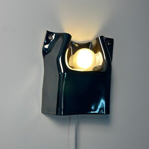 May include: A wall-mounted lamp featuring a glossy, dark, reflective ceramic body with an irregular, organic, folded shape. A single round, warm-toned light bulb is recessed within the center, casting a soft glow against the wall surface.