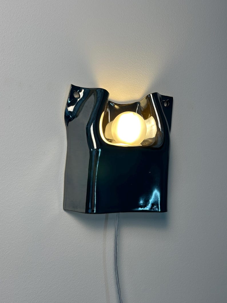 May include: A wall-mounted lamp featuring a glossy, dark, reflective ceramic body with an irregular, organic, folded shape. A single round, warm-toned light bulb is recessed within the center, casting a soft glow against the wall surface.