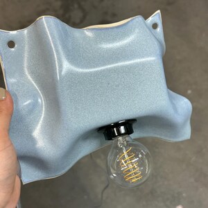 May include: A wall-mounted light fixture featuring a wavy, light blue ceramic shade with a glossy finish. The shade has two circular holes for mounting and a central black socket holding a clear glass light bulb with a visible yellow filament.