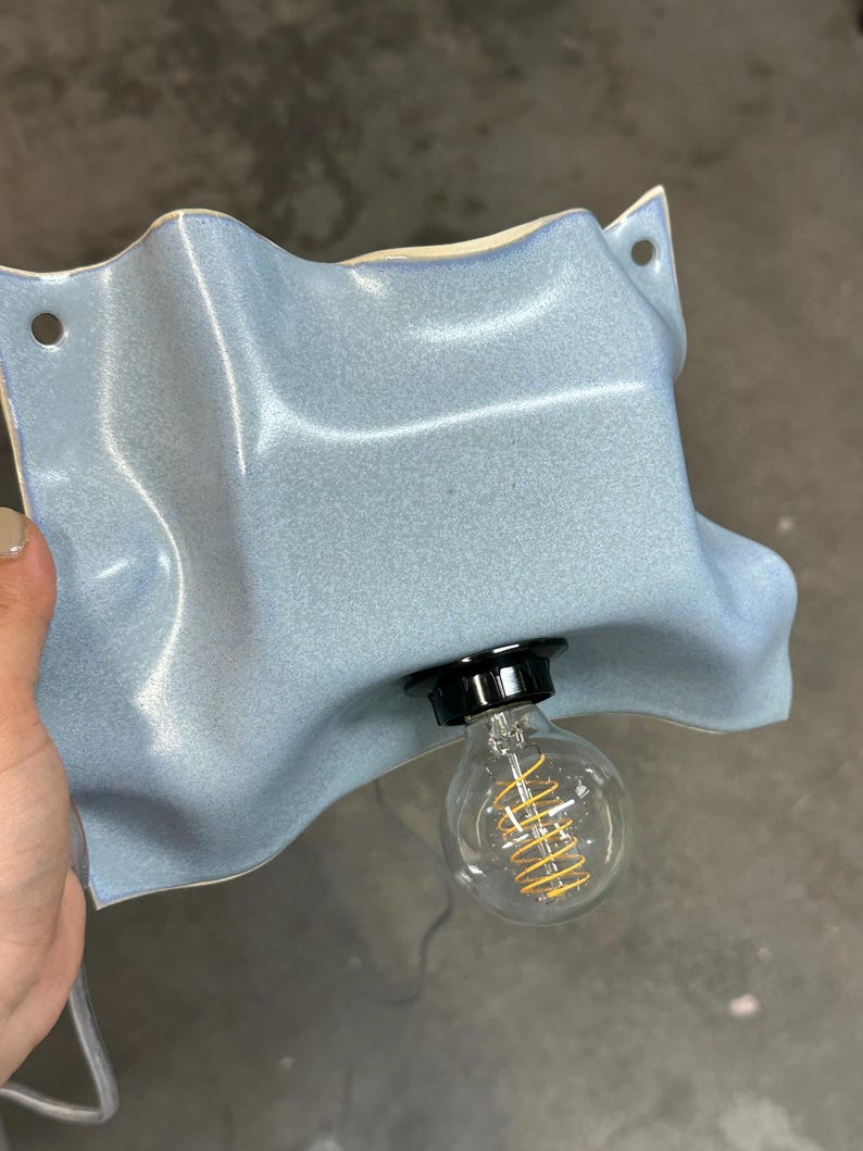 May include: A wall-mounted light fixture featuring a wavy, light blue ceramic shade with a glossy finish. The shade has two circular holes for mounting and a central black socket holding a clear glass light bulb with a visible yellow filament.