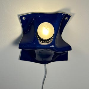 May include: A wall-mounted lamp featuring a glossy, deep blue ceramic shade with a folded, undulating form. A single round, glowing light bulb is centered within the fixture, which is secured by two visible metal screws and has a white power cord.