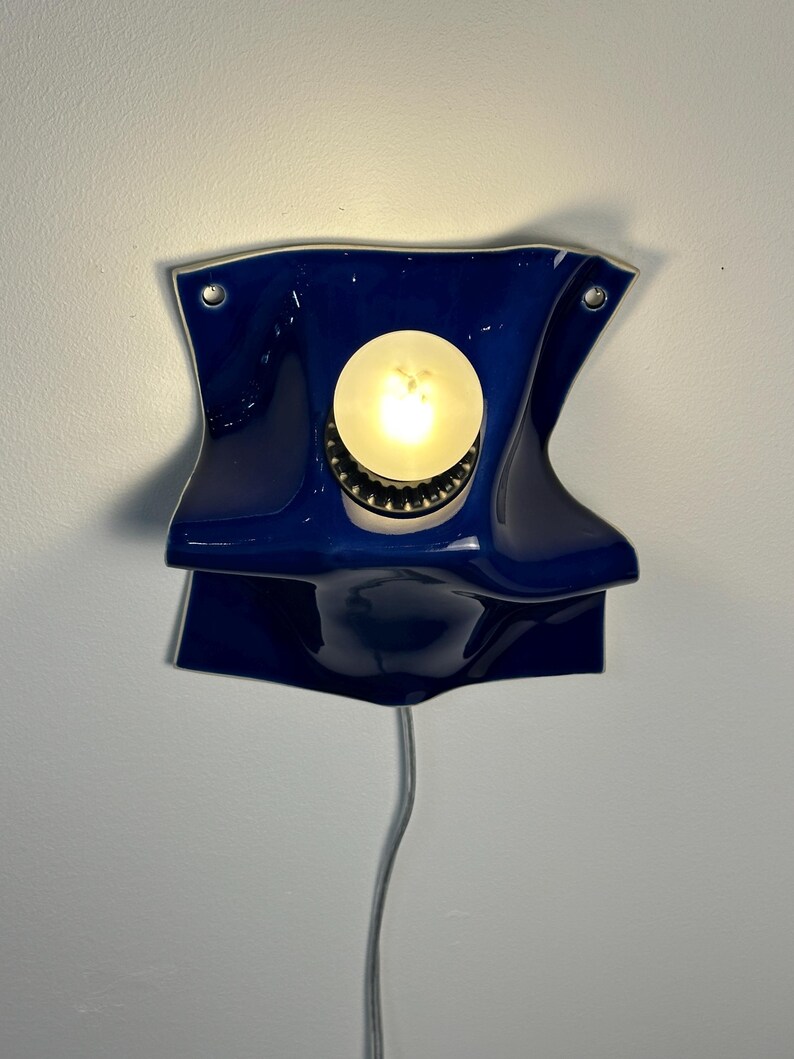 May include: A wall-mounted lamp featuring a glossy, deep blue ceramic shade with a folded, undulating form. A single round, glowing light bulb is centered within the fixture, which is secured by two visible metal screws and has a white power cord.