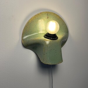 Puede incluir: Un aplique de pared de cer&aacute;mica con un esmalte verde claro y motas oscuras. La l&aacute;mpara tiene una forma curva similar a una concha e incluye un casquillo negro que sostiene una peque&ntilde;a bombilla esf&eacute;rica esmerilada brillante y un cable blanco.