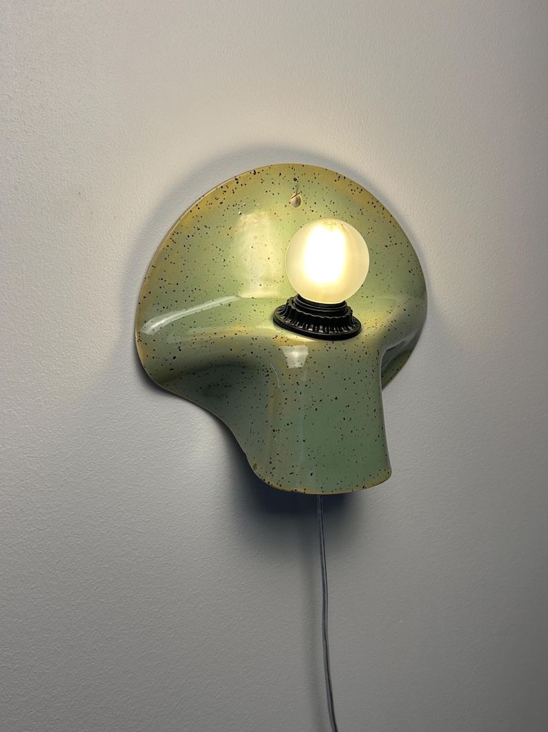 Puede incluir: Un aplique de pared de cer&aacute;mica con un esmalte verde claro y motas oscuras. La l&aacute;mpara tiene una forma curva similar a una concha e incluye un casquillo negro que sostiene una peque&ntilde;a bombilla esf&eacute;rica esmerilada brillante y un cable blanco.