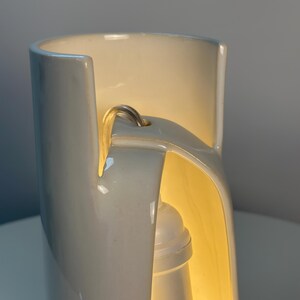 May include: A cylindrical white ceramic lamp with a smooth, glossy finish. The structure features a vertical cutout section that reveals an internal white light socket and bulb, with a power cord extending through a small hole at the top.