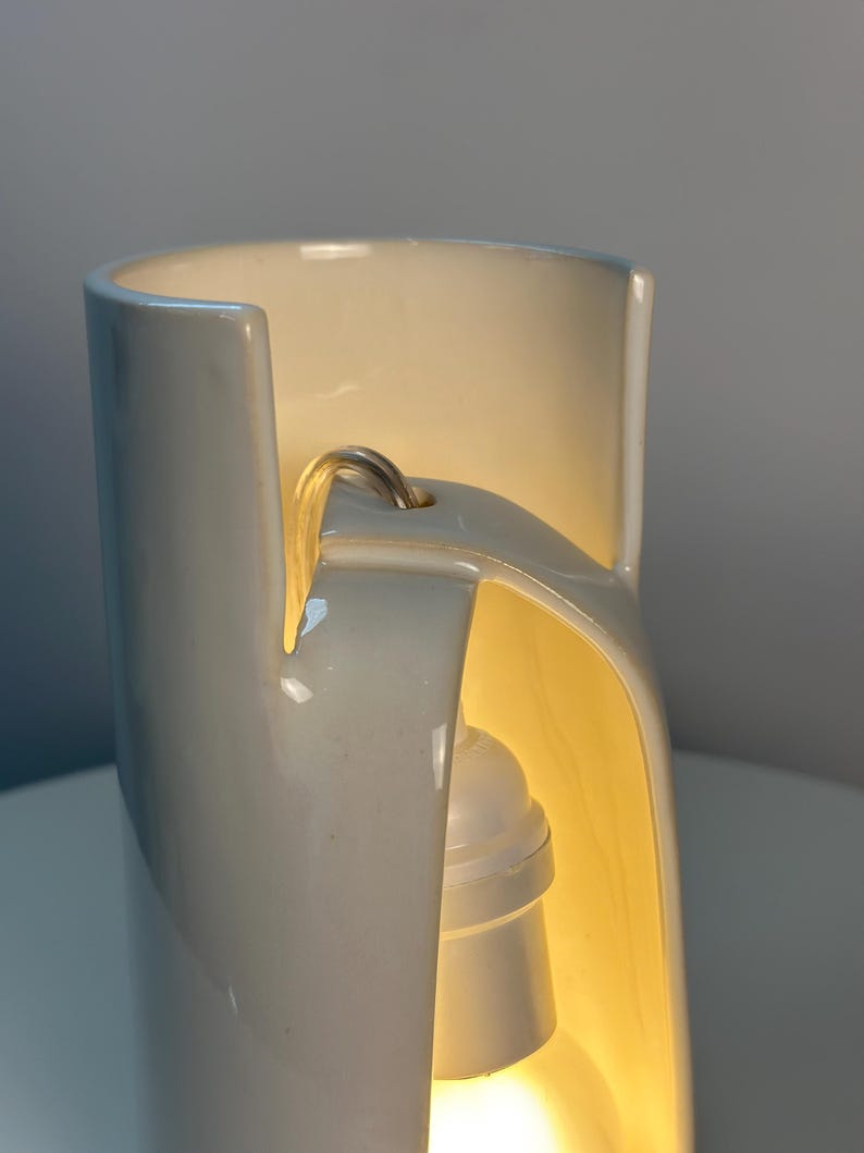 May include: A cylindrical white ceramic lamp with a smooth, glossy finish. The structure features a vertical cutout section that reveals an internal white light socket and bulb, with a power cord extending through a small hole at the top.