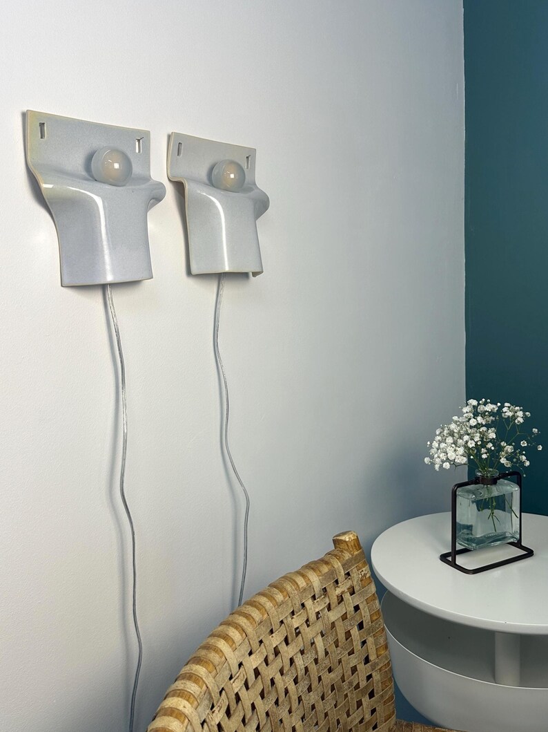 May include: Two matching wall-mounted light fixtures with a smooth, light gray ceramic finish. Each fixture features a rectangular top section with a circular cutout holding a round, frosted glass light bulb. A thin, clear power cord hangs down.