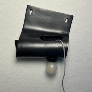Puede incluir: Una l&aacute;mpara de pared negra que presenta una l&aacute;mina rectangular curvada con dos ranuras de montaje superiores y un recorte horizontal central. Una bombilla redonda esmerilada est&aacute; unida al borde inferior mediante un cable el&eacute;ctrico transparente.
