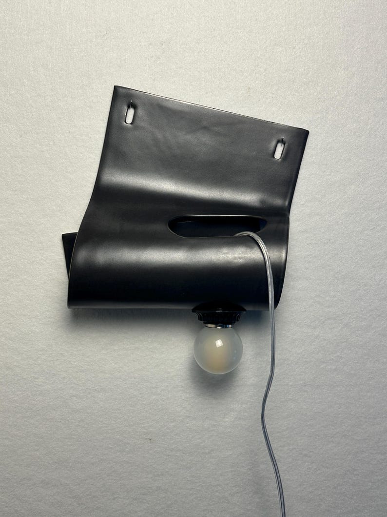 Puede incluir: Una l&aacute;mpara de pared negra que presenta una l&aacute;mina rectangular curvada con dos ranuras de montaje superiores y un recorte horizontal central. Una bombilla redonda esmerilada est&aacute; unida al borde inferior mediante un cable el&eacute;ctrico transparente.