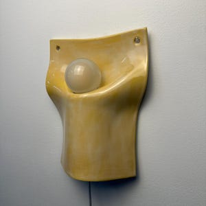 May include: A wall-mounted lamp featuring a curved, pale yellow ceramic body with a glossy finish. The fixture has two circular mounting holes at the top corners and holds a single, spherical, frosted white glass light bulb in its center.