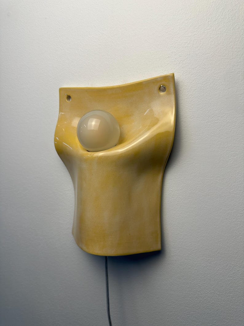May include: A wall-mounted lamp featuring a curved, pale yellow ceramic body with a glossy finish. The fixture has two circular mounting holes at the top corners and holds a single, spherical, frosted white glass light bulb in its center.
