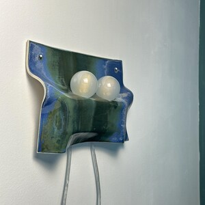 May include: A wall-mounted ceramic light fixture featuring a wavy, rectangular form with a glossy glaze in shades of deep blue and forest green. Two spherical, frosted white glass light bulbs are mounted on the front, with two clear power cords.