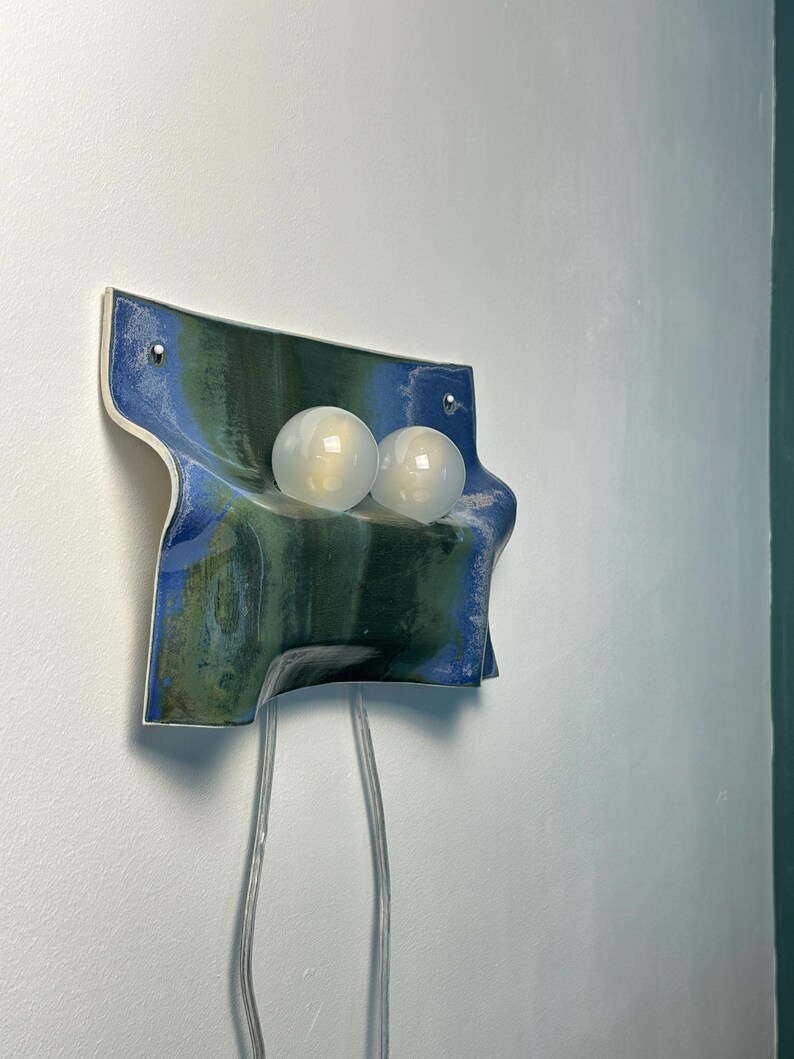 May include: A wall-mounted ceramic light fixture featuring a wavy, rectangular form with a glossy glaze in shades of deep blue and forest green. Two spherical, frosted white glass light bulbs are mounted on the front, with two clear power cords.