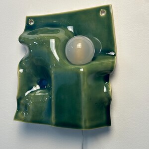 May include: A wall-mounted ceramic light fixture with a glossy, undulating green surface. The piece features a circular, frosted glass bulb integrated into the center of the wavy form. Two small holes are visible at the top for mounting purposes.