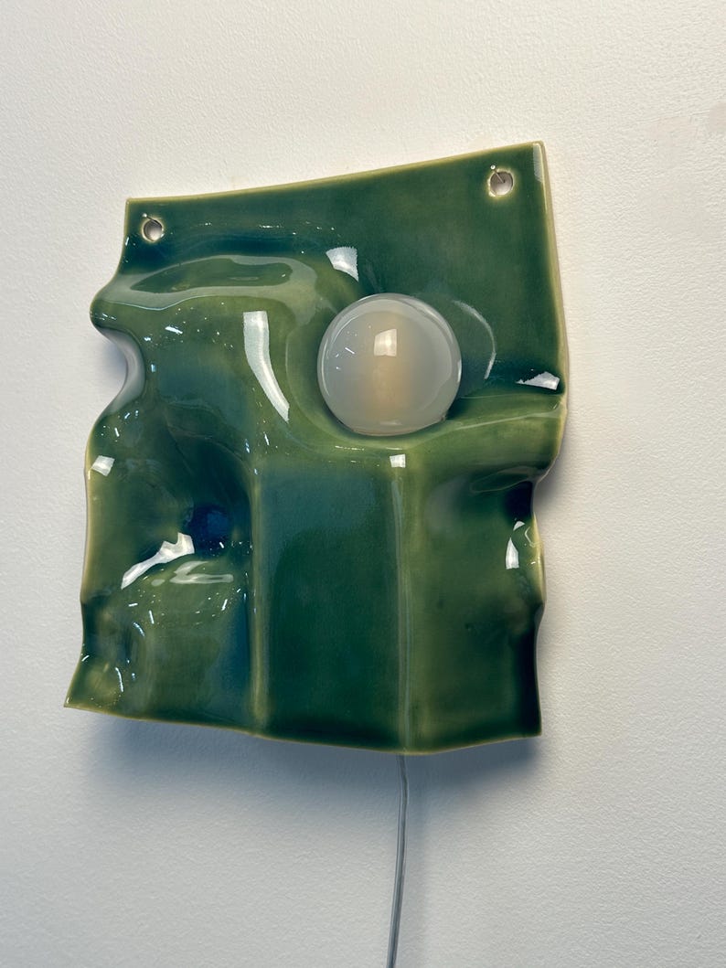 May include: A wall-mounted ceramic light fixture with a glossy, undulating green surface. The piece features a circular, frosted glass bulb integrated into the center of the wavy form. Two small holes are visible at the top for mounting purposes.