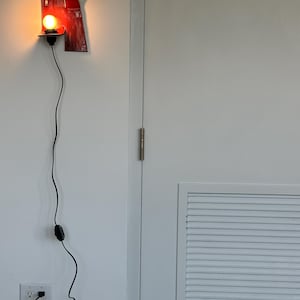 以下が含まれることがあります： 黒いコードと電球が付いた赤い壁掛けランプ。ランプはユニークなデザインの長方形をしています。コードは壁のコンセントに差し込まれています。ランプは白い壁に取り付けられています。