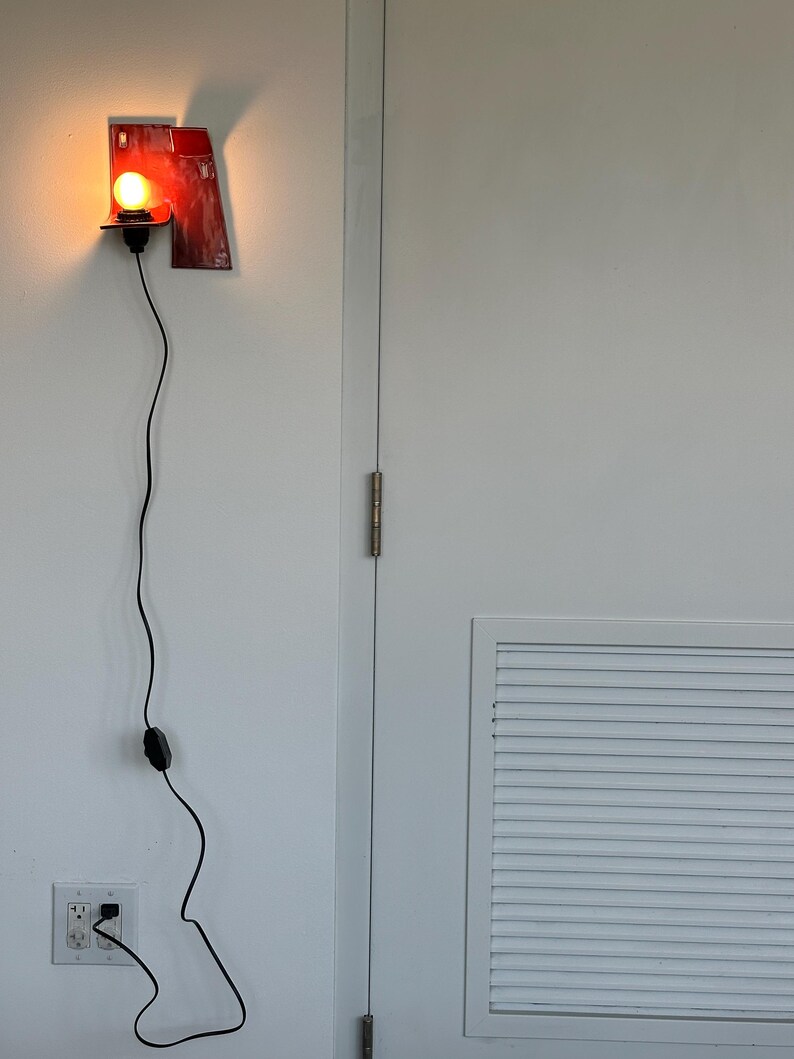 以下が含まれることがあります： 黒いコードと電球が付いた赤い壁掛けランプ。ランプはユニークなデザインの長方形をしています。コードは壁のコンセントに差し込まれています。ランプは白い壁に取り付けられています。