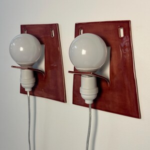 Puede incluir: Un par de apliques de pared con una placa trasera de cer&aacute;mica de color rojo oscuro brillante y forma trapezoidal. Cada l&aacute;mpara incluye un casquillo blanco, una bombilla redonda blanca y un cable el&eacute;ctrico fino de color claro hacia abajo.