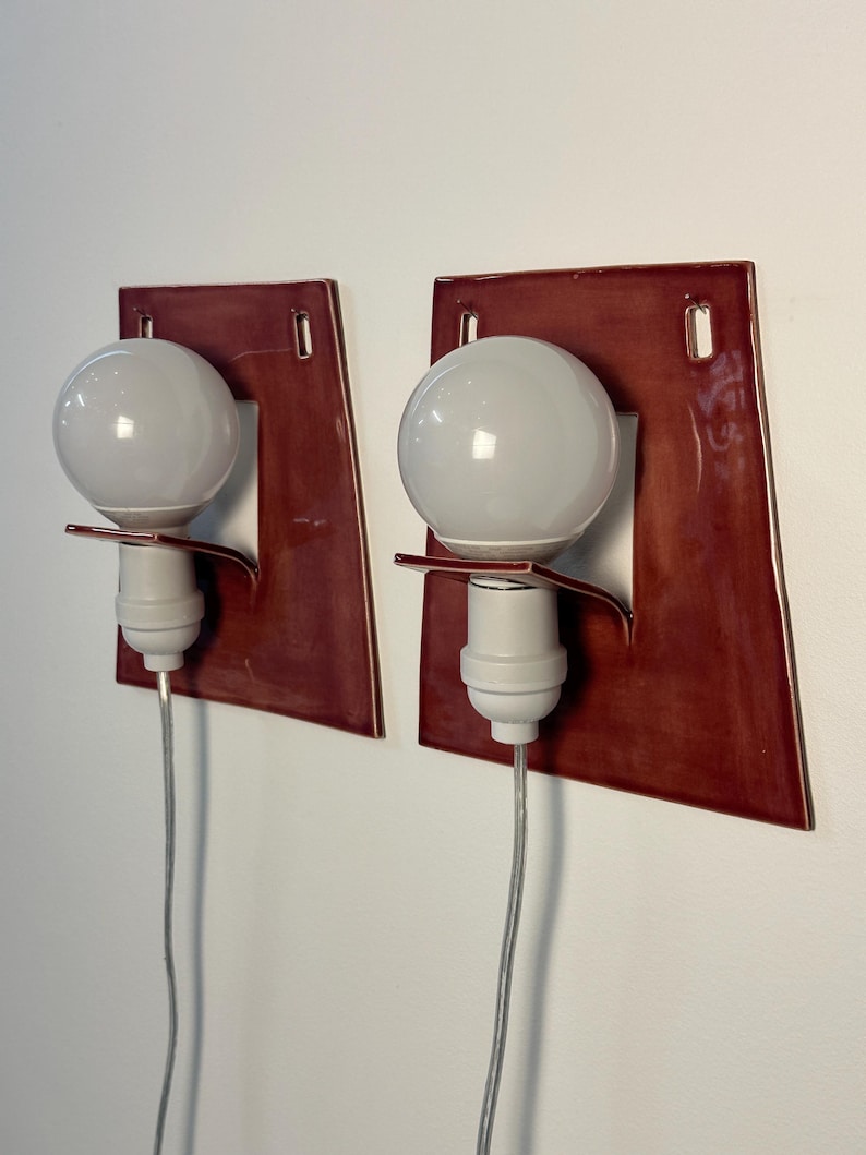 Puede incluir: Un par de apliques de pared con una placa trasera de cer&aacute;mica de color rojo oscuro brillante y forma trapezoidal. Cada l&aacute;mpara incluye un casquillo blanco, una bombilla redonda blanca y un cable el&eacute;ctrico fino de color claro hacia abajo.