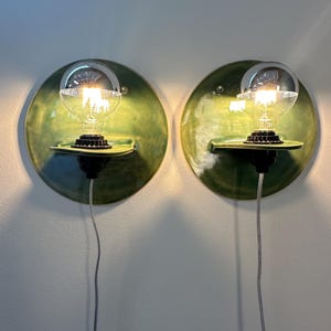 May include: A pair of circular wall sconces featuring a glossy green ceramic backplate with a small integrated shelf. Each fixture holds a clear glass light bulb with a silver-dipped top, mounted in a black socket with a thin white power cord.