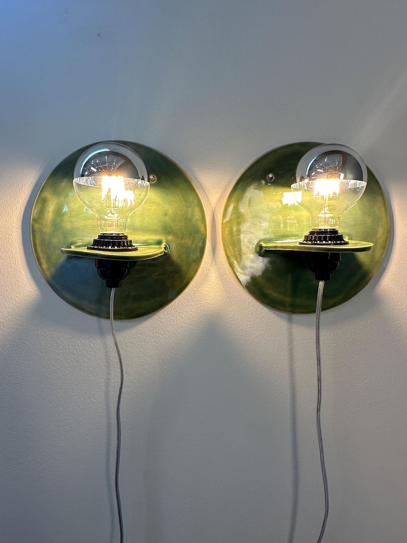 May include: A pair of circular wall sconces featuring a glossy green ceramic backplate with a small integrated shelf. Each fixture holds a clear glass light bulb with a silver-dipped top, mounted in a black socket with a thin white power cord.
