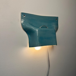 K&ouml;nnte beinhalten: Eine Wandleuchte mit einem gl&auml;nzenden, t&uuml;rkisfarbenen Keramikschirm in geschwungenem, ausgestelltem Design. Die Leuchte verf&uuml;gt &uuml;ber einen kleinen Metallgriff an der Oberseite, zwei Befestigungsl&ouml;cher an den oberen Ecken und eine leuchtende Gl&uuml;hbirne.