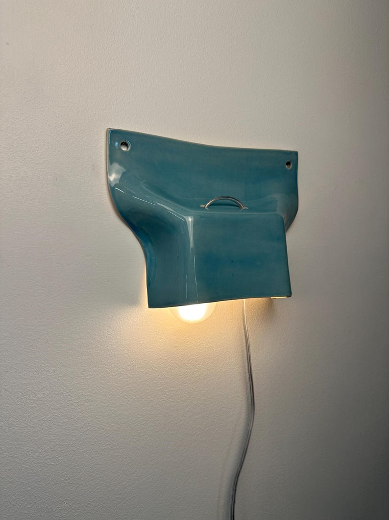 K&ouml;nnte beinhalten: Eine Wandleuchte mit einem gl&auml;nzenden, t&uuml;rkisfarbenen Keramikschirm in geschwungenem, ausgestelltem Design. Die Leuchte verf&uuml;gt &uuml;ber einen kleinen Metallgriff an der Oberseite, zwei Befestigungsl&ouml;cher an den oberen Ecken und eine leuchtende Gl&uuml;hbirne.