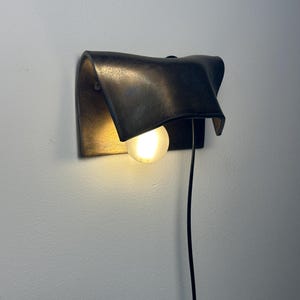 Pode incluir: Um candeeiro de parede com um abajur met&aacute;lico escuro, curvo e com acabamento texturizado. Uma l&acirc;mpada redonda e brilhante est&aacute; posicionada sob o abajur, emitindo uma luz quente. Um fio de alimenta&ccedil;&atilde;o preto fino pende verticalmente.
