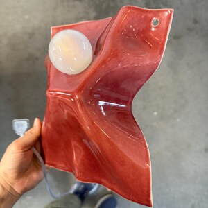 Puede incluir: Una l&aacute;mpara de pared con una pantalla de cer&aacute;mica de color rojo intenso brillante con una silueta irregular, plegada y ondulada. El accesorio incluye una bombilla redonda de cristal blanco y un peque&ntilde;o orificio circular en la esquina superior.