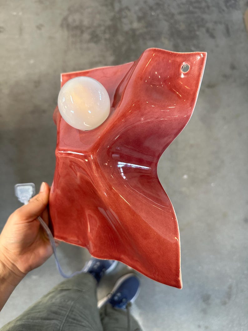 Puede incluir: Una l&aacute;mpara de pared con una pantalla de cer&aacute;mica de color rojo intenso brillante con una silueta irregular, plegada y ondulada. El accesorio incluye una bombilla redonda de cristal blanco y un peque&ntilde;o orificio circular en la esquina superior.
