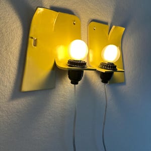 May include: Two yellow wall-mounted sconces featuring a flat, angular metal backplate with a protruding shelf. Each fixture holds a single round, glowing white light bulb in a black socket, with a thin, clear power cord extending downward.