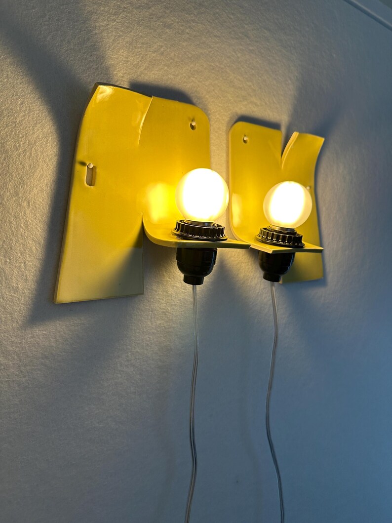 May include: Two yellow wall-mounted sconces featuring a flat, angular metal backplate with a protruding shelf. Each fixture holds a single round, glowing white light bulb in a black socket, with a thin, clear power cord extending downward.