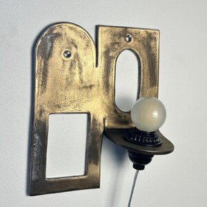 K&ouml;nnte beinhalten: Eine Wandleuchte mit einer flachen, messingfarbenen Metallplatte, die einen bogenf&ouml;rmigen Ausschnitt und eine rechteckige &Ouml;ffnung aufweist. Eine kleine, runde, mattwei&szlig;e Gl&uuml;hbirne sitzt in einer schwarzen Fassung auf einer Ablage.