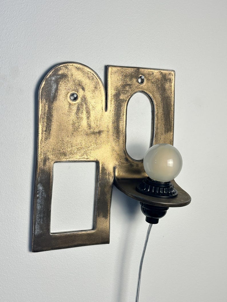 K&ouml;nnte beinhalten: Eine Wandleuchte mit einer flachen, messingfarbenen Metallplatte, die einen bogenf&ouml;rmigen Ausschnitt und eine rechteckige &Ouml;ffnung aufweist. Eine kleine, runde, mattwei&szlig;e Gl&uuml;hbirne sitzt in einer schwarzen Fassung auf einer Ablage.